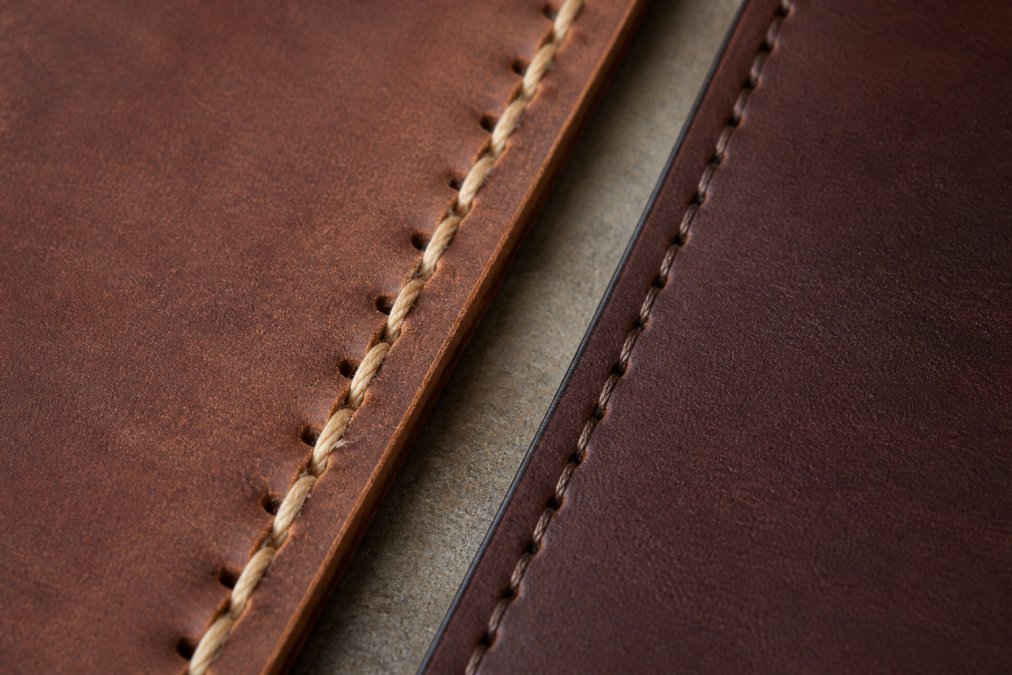 Close-up of leather seam showing individual hand-stitched saddle stitches with visible needle holes and thread, contrasted with uniform machine stitching on parallel leather edge in natural daylight