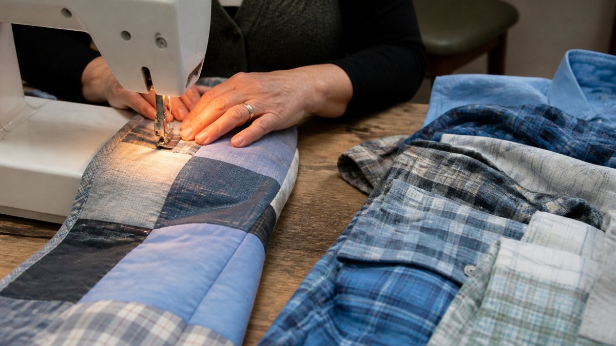 Close-up of a quilter's hands guiding fabric under a sewing machine needle, with a patchwork of a loved one's shirts laid out on a wooden worktable nearby