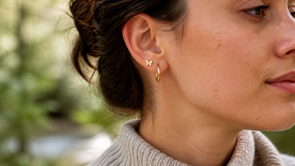 Close-up of a woman's ear wearing small gold hoop earrings and a delicate butterfly stud, natural daylight, casual knit sweater, relaxed expression