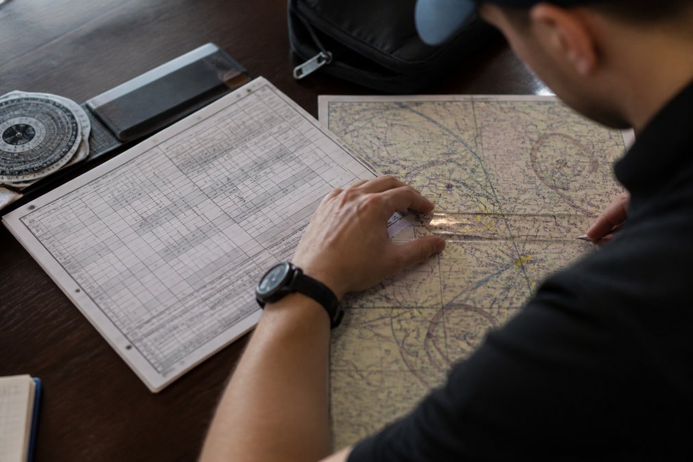 Dispatcher or student pilot with navigation log worksheet, aeronautical chart, and flight planning tools spread across a desk, using a ruler or plotter to measure distances between waypoints on the chart
