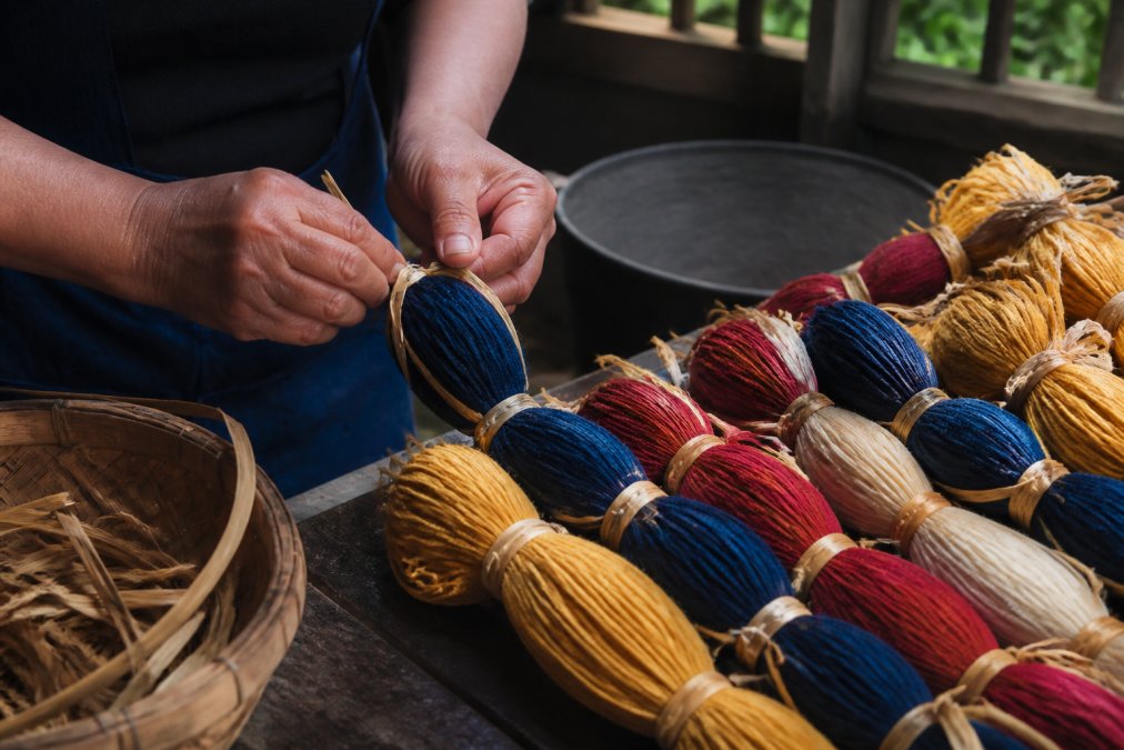 Close-up of hands carefully tying sections of yarn with natural fiber raffia before dyeing, showing the intricate binding technique with multiple colors of yarn in a traditional workspace with natural window lighting