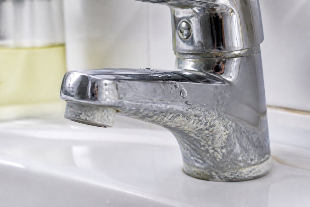 Close-up of a chrome faucet with thick white limescale crust forming around the base and spout, water droplets beading on the mineral deposits, bright bathroom lighting