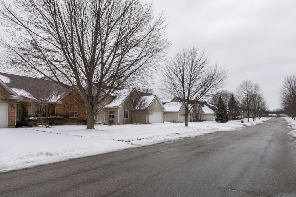 Empty suburban street lined with homes during winter with snow on ground and bare trees, showing quiet neighborhood with few people or activity