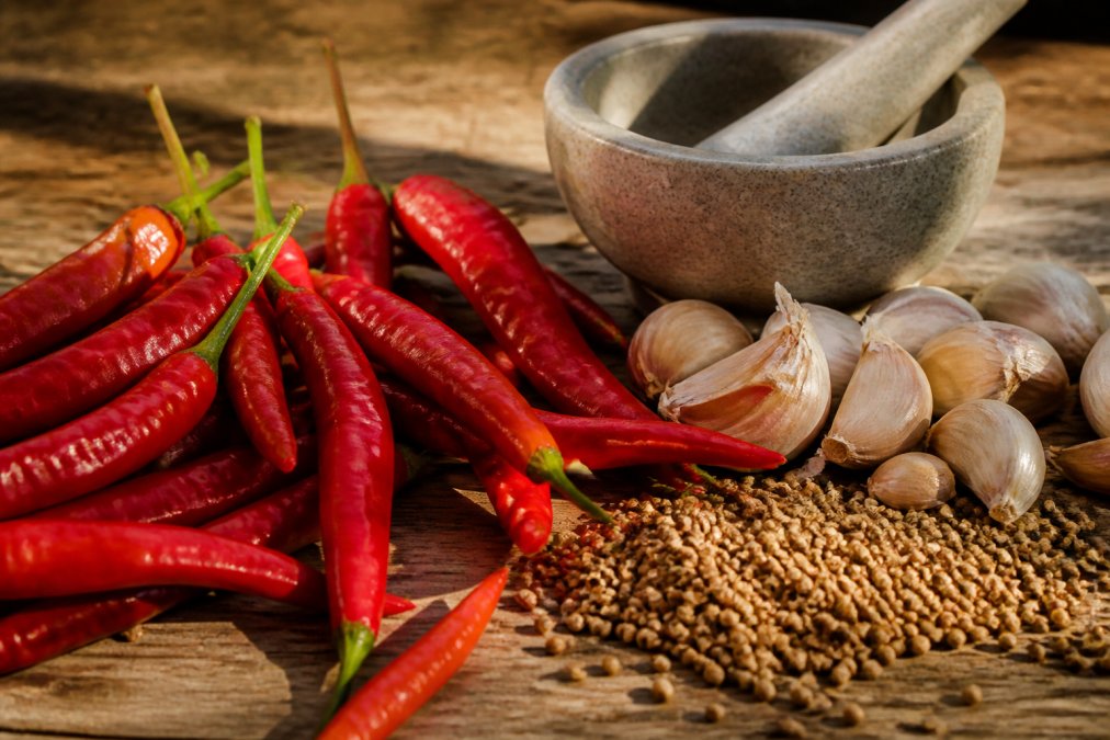 Close-up of fresh red chili peppers, garlic cloves, and whole coriander seeds arranged on a rustic wooden surface with natural sunlight, with a mortar and pestle visible in the background
