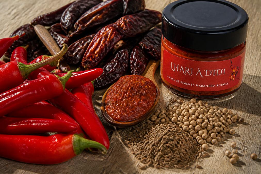 Gros plan sur des piments rouges frais brillants et des piments séchés fumés foncés, avec des graines de carvi, de coriandre et de cumin dispersées à côté d'une cuillère en bois contenant une pâte harissa rougeâtre granuleuse, sur une surface rustique en bois clair éclairée par la lumière naturelle