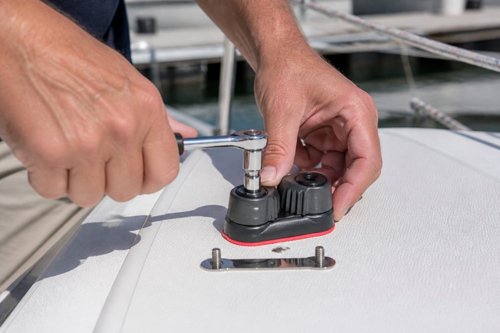Hands installing Harken cleat onto deck with socket wrench, stainless steel fasteners visible, showing proper alignment and fastener placement with deck backing plate