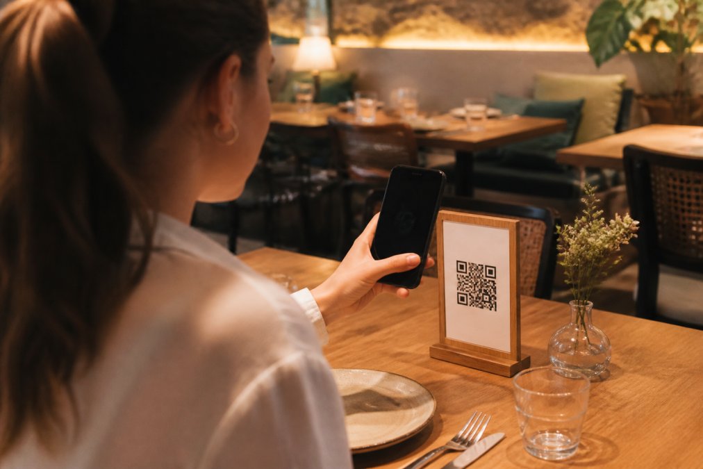 Cliente en una mesa de restaurante escaneando un código QR en un soporte de mesa con su smartphone, sala con iluminación cálida y ambiente acogedor al fondo