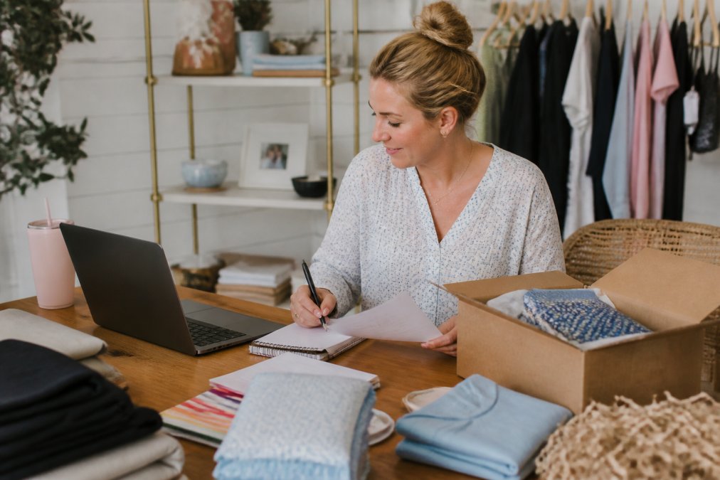 Small business owner or artisan working at a laptop with inventory items, packaging materials, and order notes organized on desk, managing multi-channel sales setup