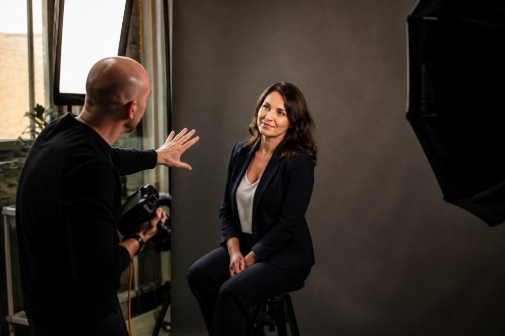 Professional photographer directing a client during a headshot session in a well-lit studio, adjusting lighting softboxes and providing posture guidance while the client sits confidently against a neutral grey backdrop