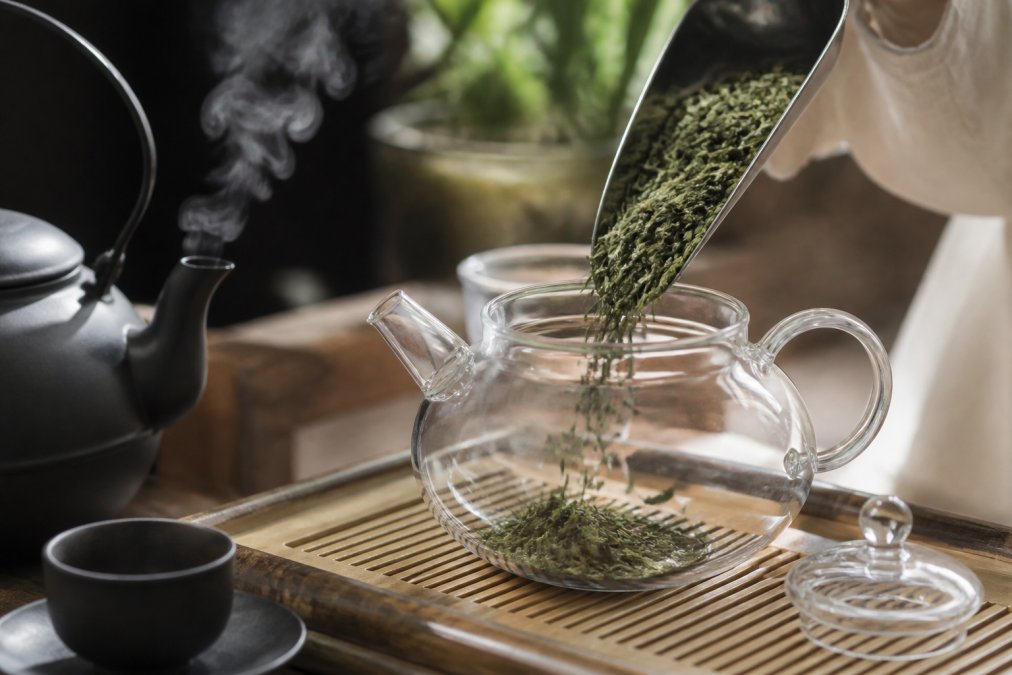 Vibrant high quality green tea leaves being poured into a glass teapot with rising steam
