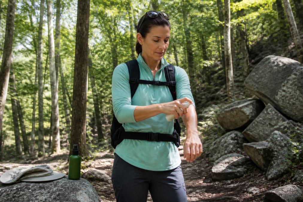 A hiker in a wooded area applying insect repellent spray to their forearm, bottle resting on a nearby rock, wearing hiking clothes and a backpack in dappled afternoon light