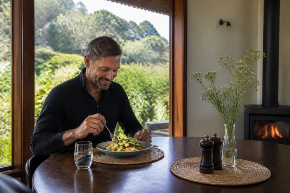 Guest enjoying organic meal at intimate table in retreat dining space with natural light streaming through windows overlooking native gardens