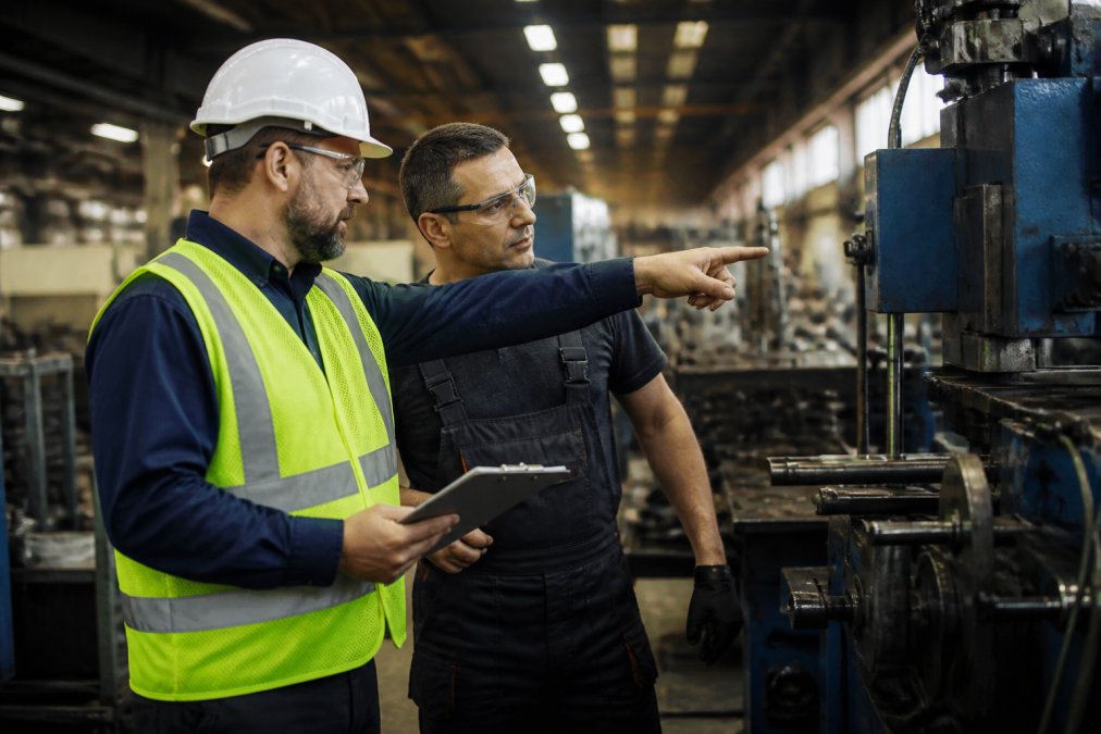 A safety professional in a hard hat and high-visibility vest reviewing a clipboard with a plant manager on a manufacturing floor, pointing at equipment