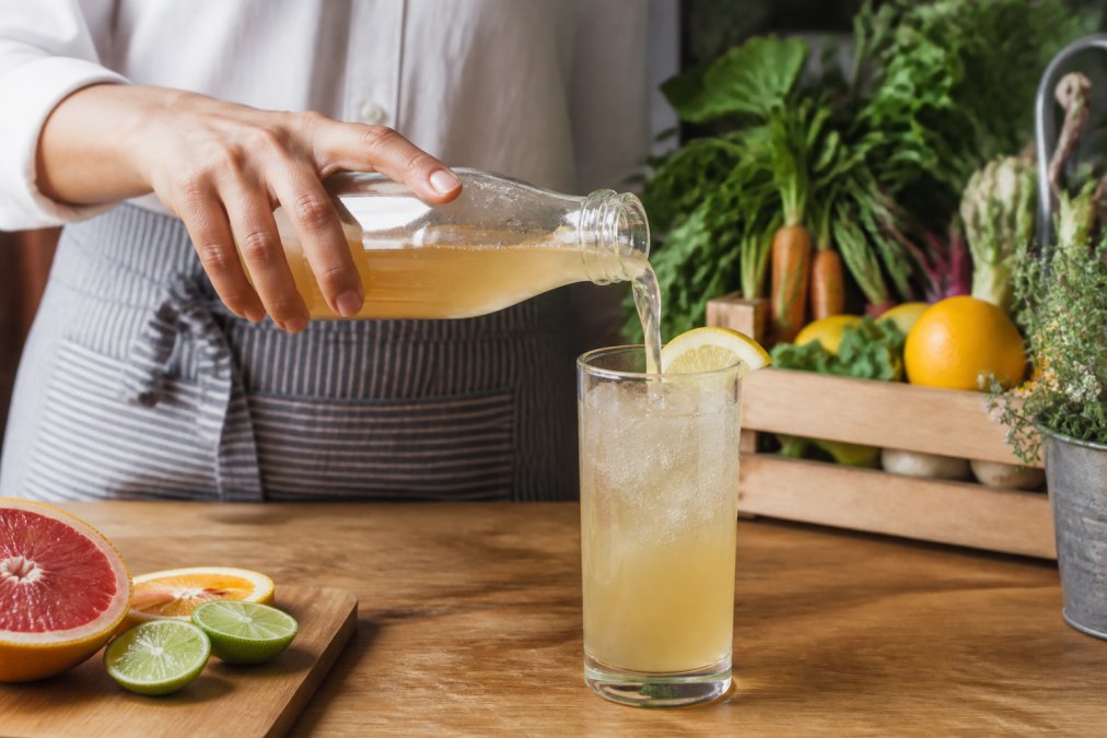 A person pouring raw kombucha from a refrigerated glass bottle into a tall glass with ice, fresh citrus and a wooden crate of farmers market produce visible on the counter behind