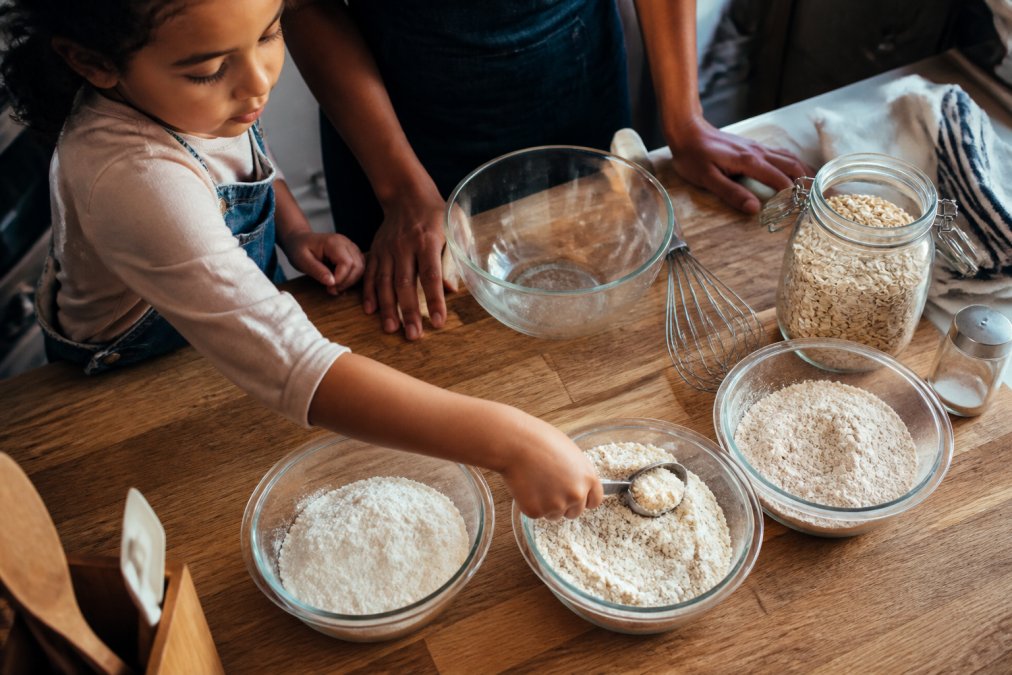 A close-up overhead shot of a wooden kitchen counter with small glass bowls holding oat flour, whole wheat flour, and a gluten-free blend, a child's hand reaching in to scoop, warm afternoon light
