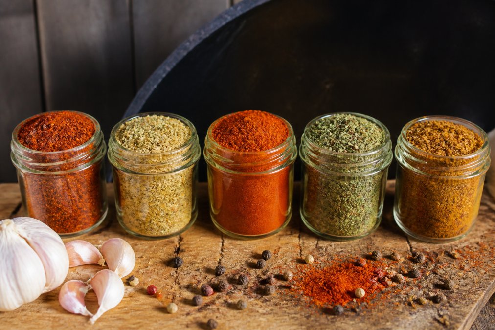 Close-up of five colorful spice rubs in glass jars arranged on a wooden surface with fresh garlic, peppercorns, and smoked paprika scattered nearby
