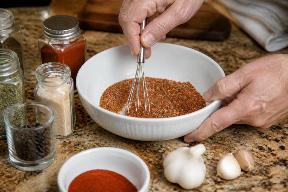 Hands mixing dry rub ingredients in a white bowl on a kitchen counter, with individual spice containers and fresh garlic, peppercorns, and smoked paprika visible in the background
