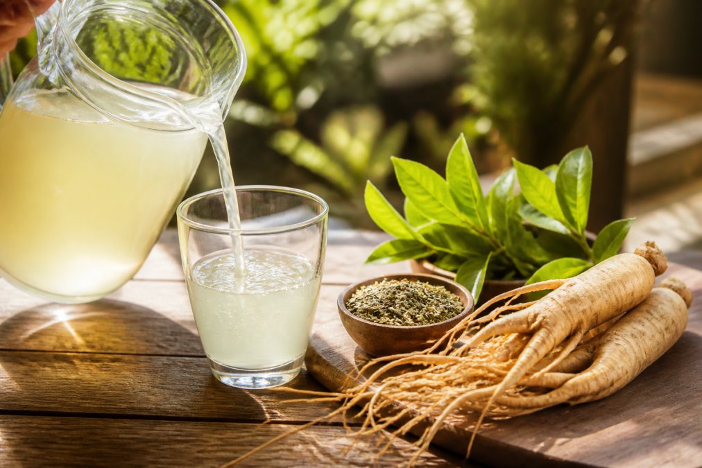 A clear glass pitcher of electrolyte drink being poured next to fresh green tea leaves and ginseng root on a sunlit wooden table