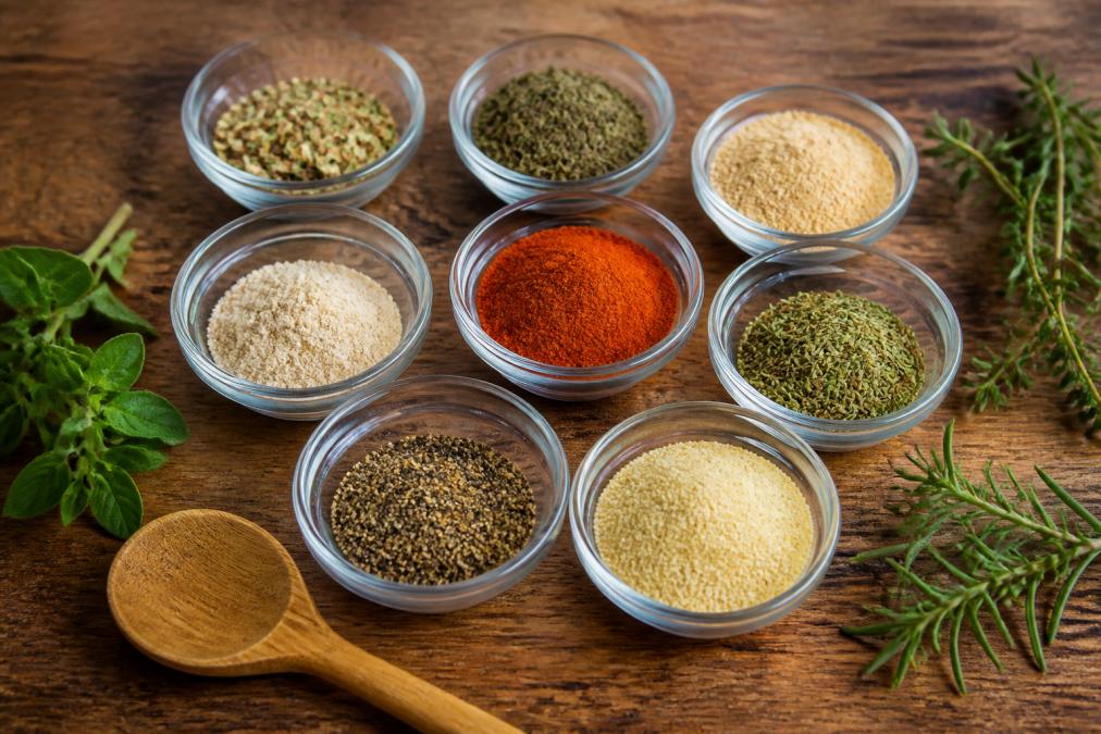 Overhead shot of small glass bowls filled with colorful salt-free spices and herbs, with a wooden spoon and fresh herbs on a rustic kitchen counter