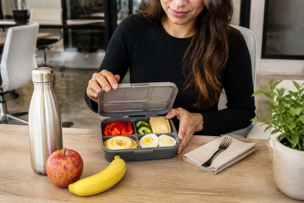A woman at a clean, modern office desk opening a divided bento box filled with hummus, sliced red bell peppers and cucumbers, hard-boiled eggs, and whole grain crackers, with a stainless steel reusable water bottle and fresh fruit on the side