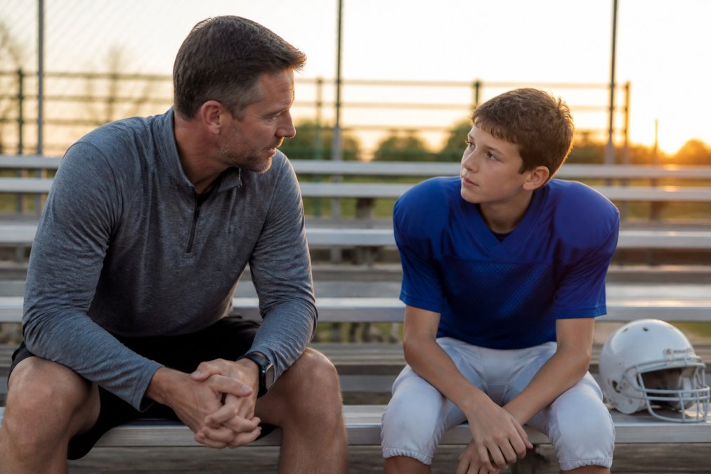 Parent and young athlete sitting together on a sideline bench after a game, having a calm conversation with relaxed body language and attentive eye contact, natural late-afternoon lighting