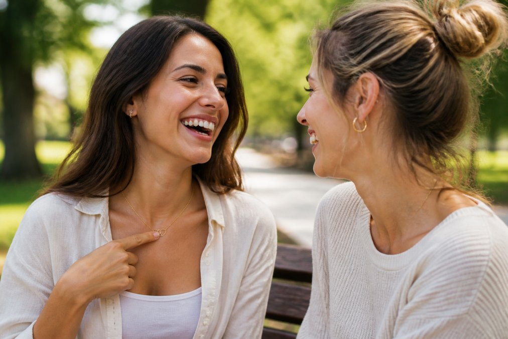 Close-up of two women laughing together on a sunlit park bench; one gestures to the delicate interlocking hearts pendant resting on her collarbone, the gold finish catching the light