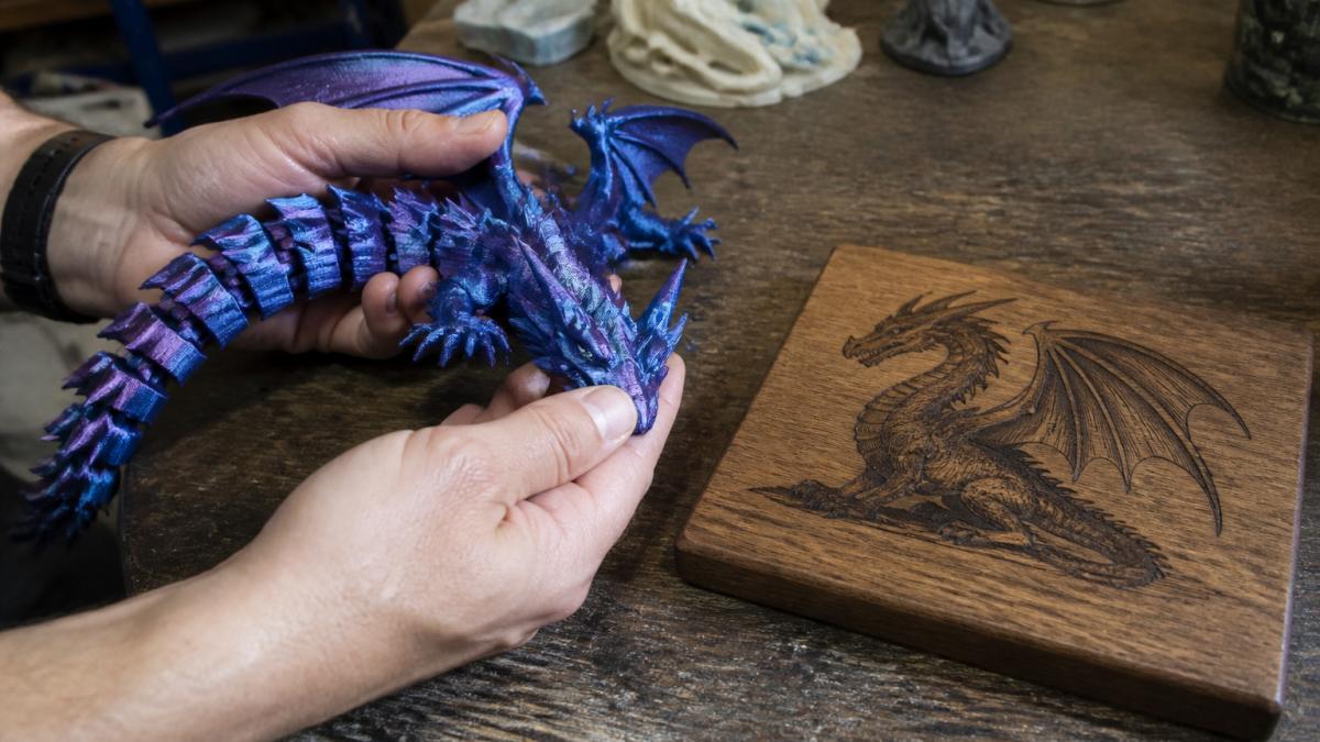 Close-up of hands holding a 3D-printed articulated dragon with visible hinged segments and a smooth filament finish, beside a laser-engraved wooden plaque on a workbench