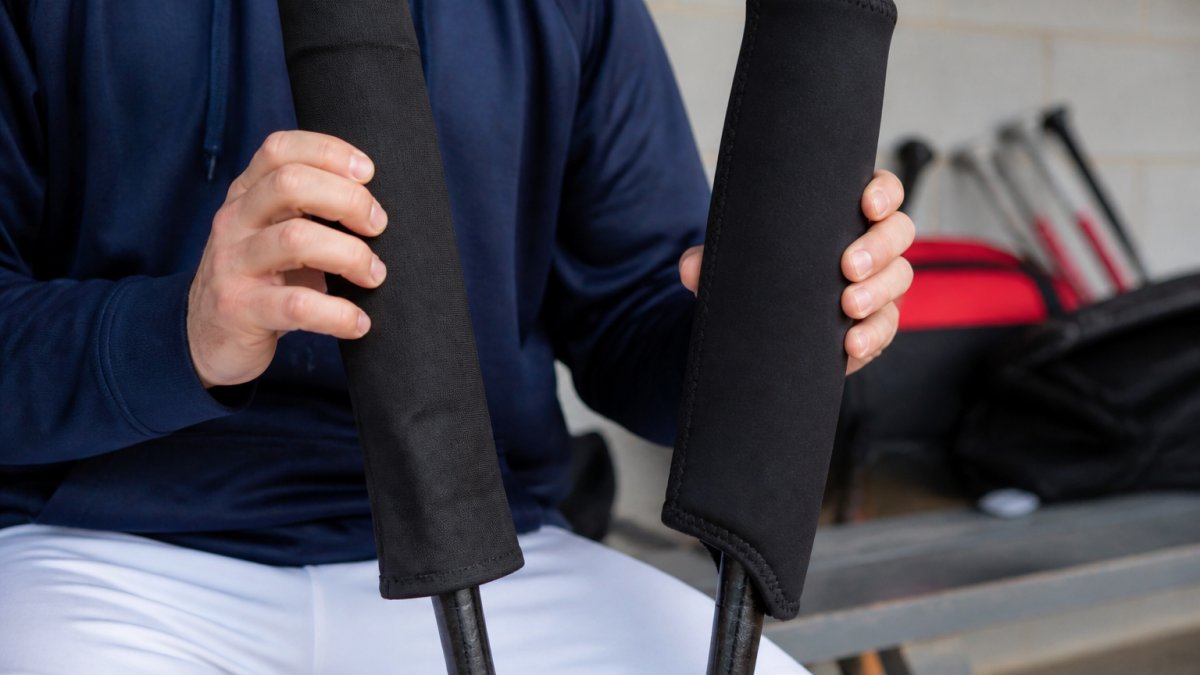 Close-up of a player in a dugout holding a heated bat sleeve on one bat and an insulated neoprene cover on another, showing the visible difference in thickness and construction between the two