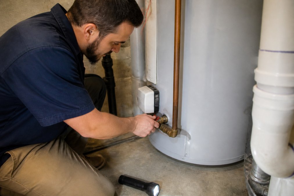 A homeowner in work clothes turning the shut-off valve on a residential water heater in a basement, with a flashlight nearby