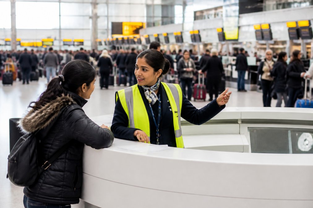Airport terminal staff member in a high-visibility vest assisting a passenger at an information desk, with a busy departure lounge in the background