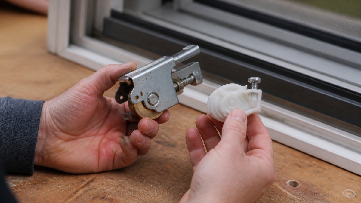 Close-up of a homeowner's hands comparing a heavy-duty metal sliding screen door roller assembly with a standard plastic roller on a workbench, with a sliding screen door frame visible in the background