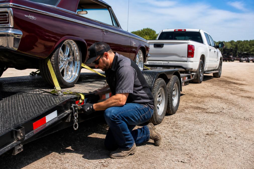 A person in work clothes kneeling beside a flatbed car hauler trailer, checking safety chains and tie-down straps attached to a classic car, with a pickup truck hitched in the background at an outdoor rental lot on a clear day