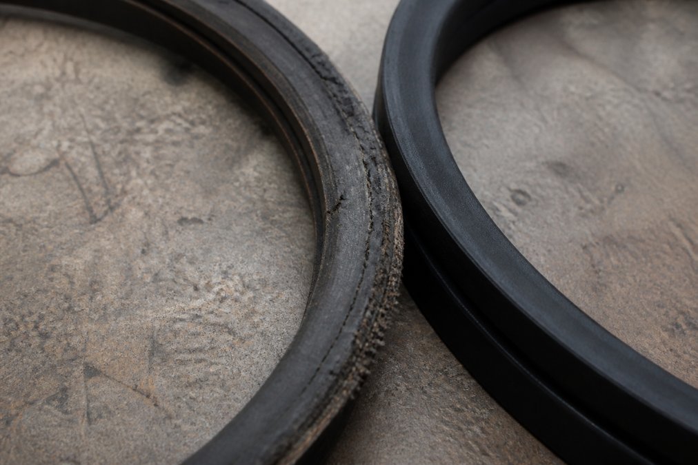 Close-up of a worn mower belt showing visible cracks, fraying edges, and glazing on the rubber surface, positioned next to a new unworn belt for comparison