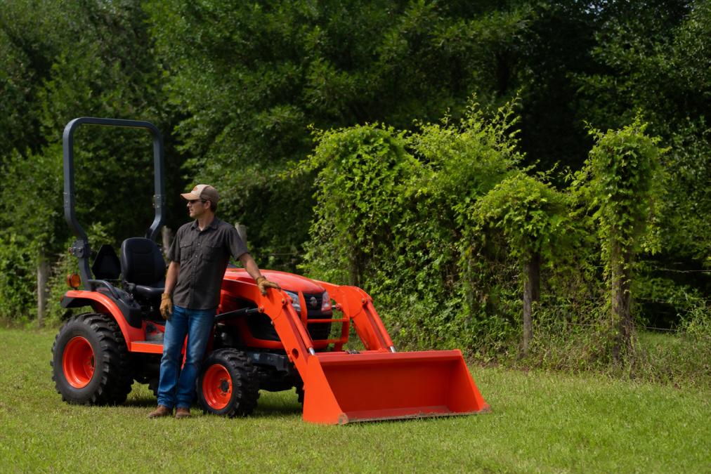 A person standing by a compact utility tractor with a front-end loader, an example of heavy equipment for small acreage.