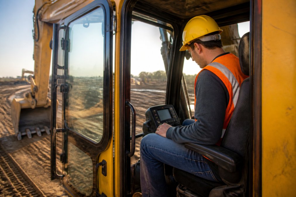 A new operator in a high-visibility orange vest and yellow hard hat sitting in the cab of a large yellow excavator on an active construction site, reviewing the control panel before operation, morning light casting long shadows across the dirt jobsite