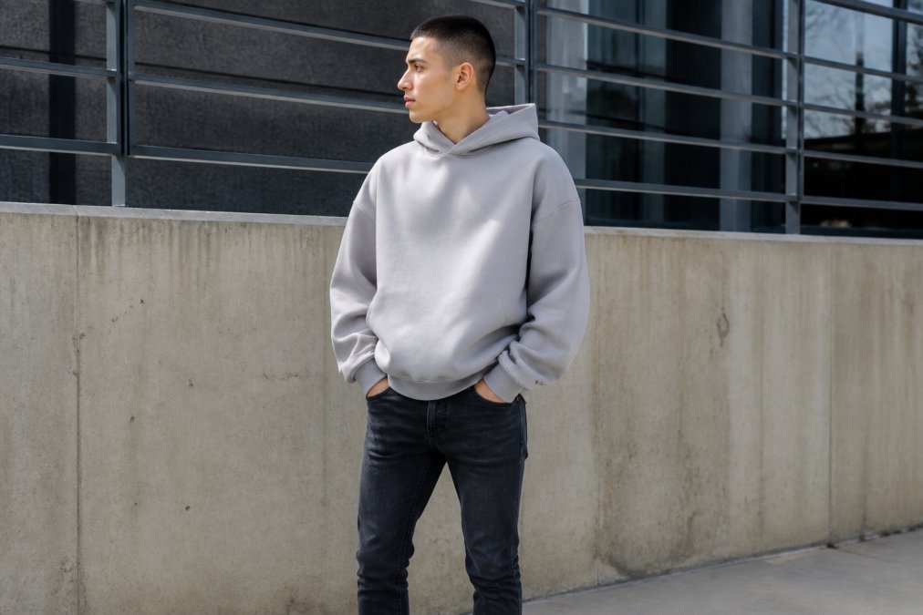 Person wearing an oversized heavyweight hoodie with dropped shoulders paired with fitted dark denim, standing outdoors in natural daylight with neutral urban background to showcase the relaxed silhouette and structured streetwear aesthetic