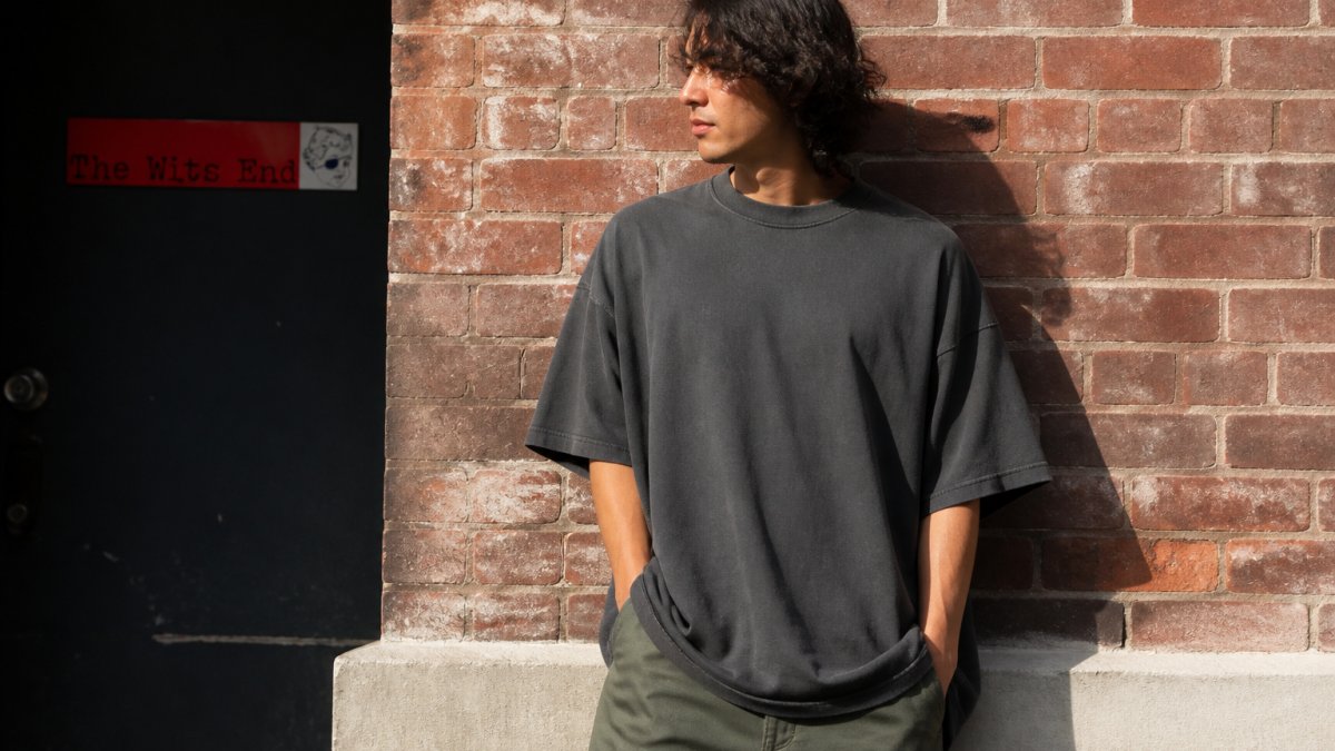 A person wearing an oversized heavyweight graphic tee standing against a brick wall in soft afternoon light, showing the structured drape and boxy silhouette of the fabric