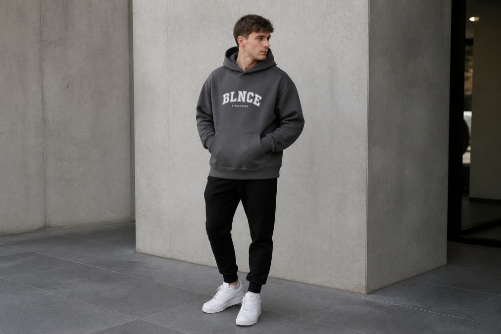 Young adult wearing an oversized heavyweight 350GSM cotton hoodie in charcoal styled with fitted black joggers and white trainers, photographed in natural daylight showing the relaxed boxy silhouette and how the structured fabric drapes across the shoulders and torso