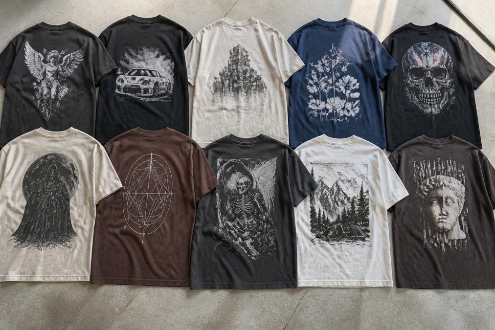 Flat-lay arrangement of multiple heavyweight graphic t-shirts displayed to show different designs, fit profiles, and color variations side by side on neutral background