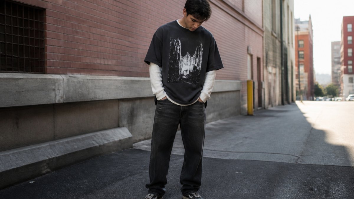 Person wearing an oversized heavyweight graphic tee layered over a long-sleeve thermal shirt, styled with worn dark denim and vintage sneakers, standing in an urban alley with brick walls and natural afternoon lighting