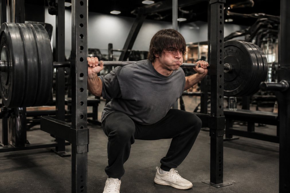 Athlete performing heavy barbell squat wearing oversized heavyweight cotton tee, demonstrating fabric structure and support during intense compound lift in modern gym with bright overhead lighting