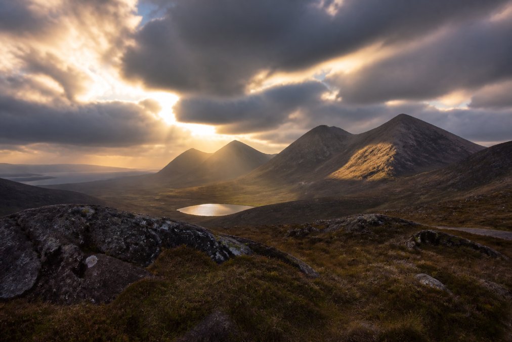 Dramatic Highland mountain landscape with moody sky, deep shadows in valleys, and golden light breaking through clouds over towering peaks and rocky terrain