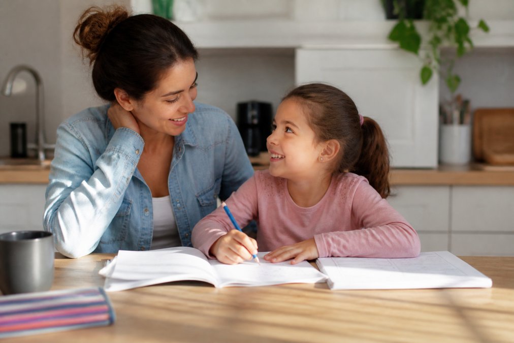 Parent and elementary-aged child sitting together at kitchen table with math homework, child smiling, natural window lighting streaming across the table, relaxed and supportive atmosphere