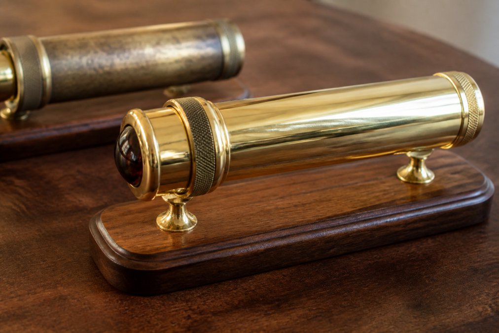Close-up of a handcrafted brass kaleidoscope with a polished barrel resting on a wooden display base on a dark walnut table, warm side lighting catching the metal and a second vintage scope slightly out of focus behind it