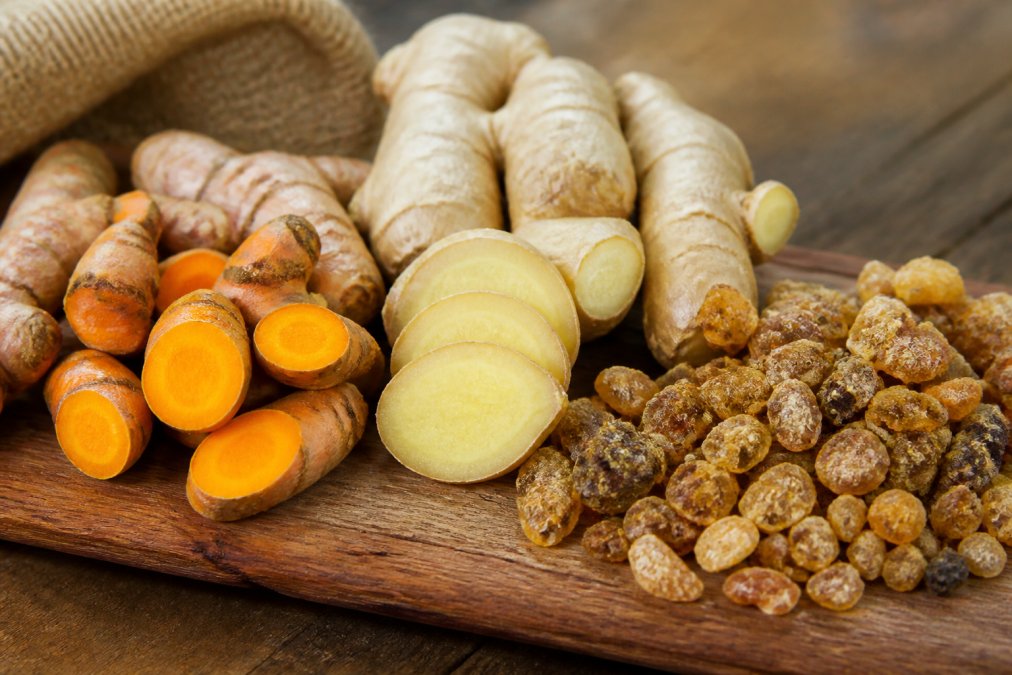 Close-up of fresh turmeric root, ginger root, and dried boswellia arranged on a wooden surface with warm natural lighting, showcasing the golden and brown tones of these dried herbal remedies