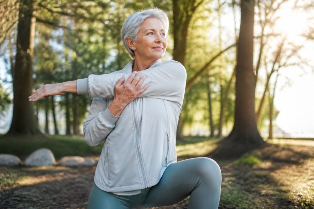 An older adult doing gentle stretching exercises outdoors in natural morning sunlight, demonstrating joint mobility and active wellness with trees visible in the background