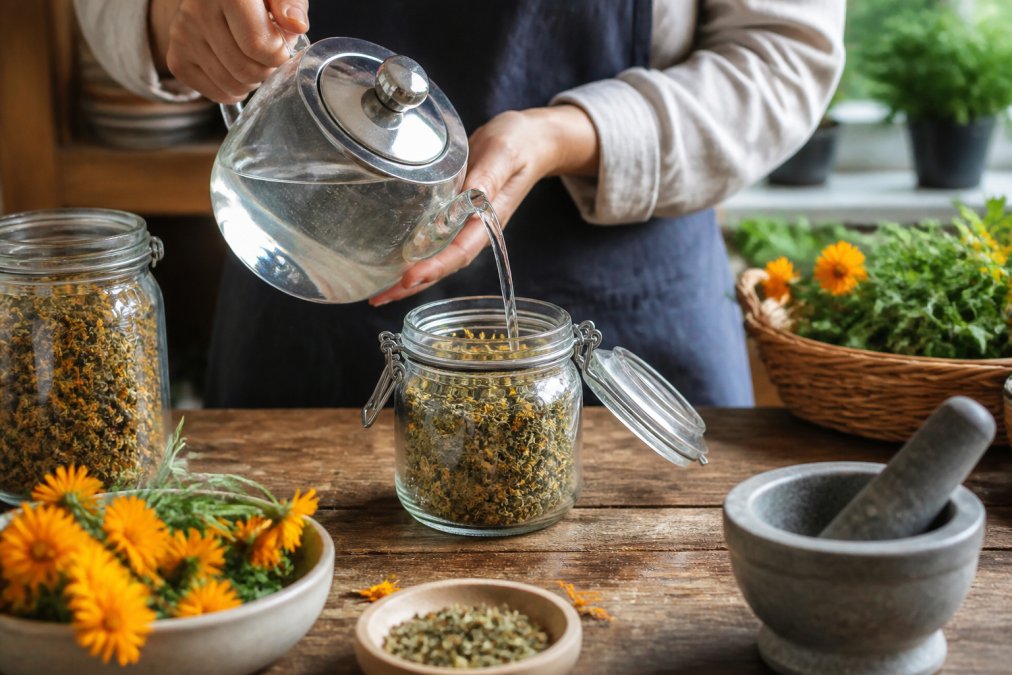 Close-up of hands pouring hot water over dried herbs in a glass jar on a wooden kitchen counter, with fresh calendula flowers and a mortar and pestle nearby in natural morning light