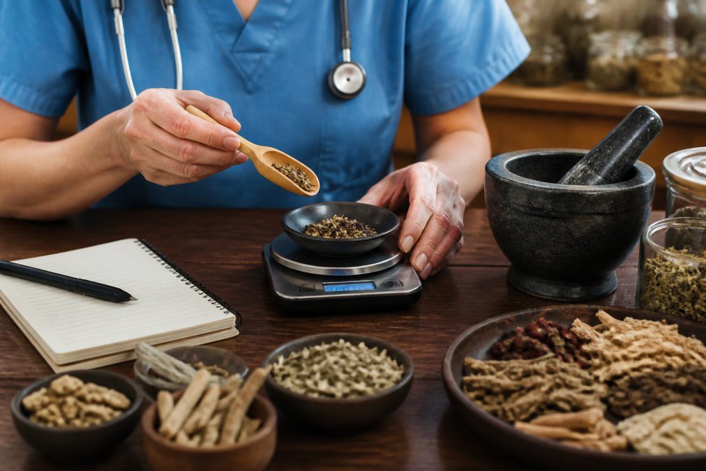 Close-up of a registered nurse's hands measuring dried adaptogenic herbs on a small kitchen scale beside a mortar and pestle, with handwritten notes on a notepad nearby