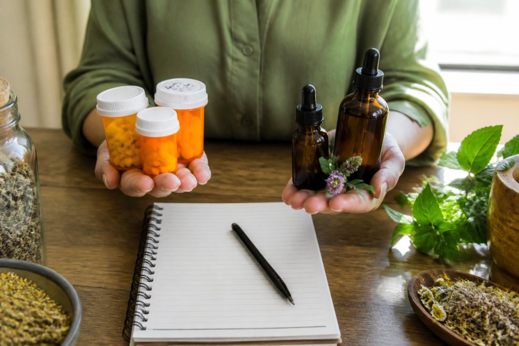 Hands holding prescription bottles and herbal tinctures side by side on desk with notebook and pen for tracking interactions, natural morning light