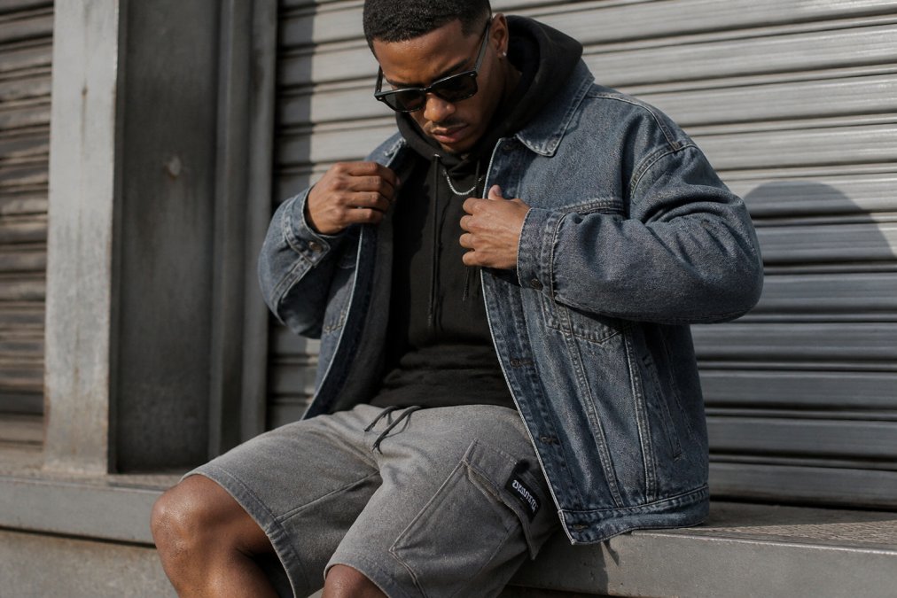Close-up detailshot van gelaagde streetwear outfit: oversized denim jacket over hoodie met cargo shorts, zichtbare textuur van stoffen en nauwkeurige pasvorm in natuurlijk licht